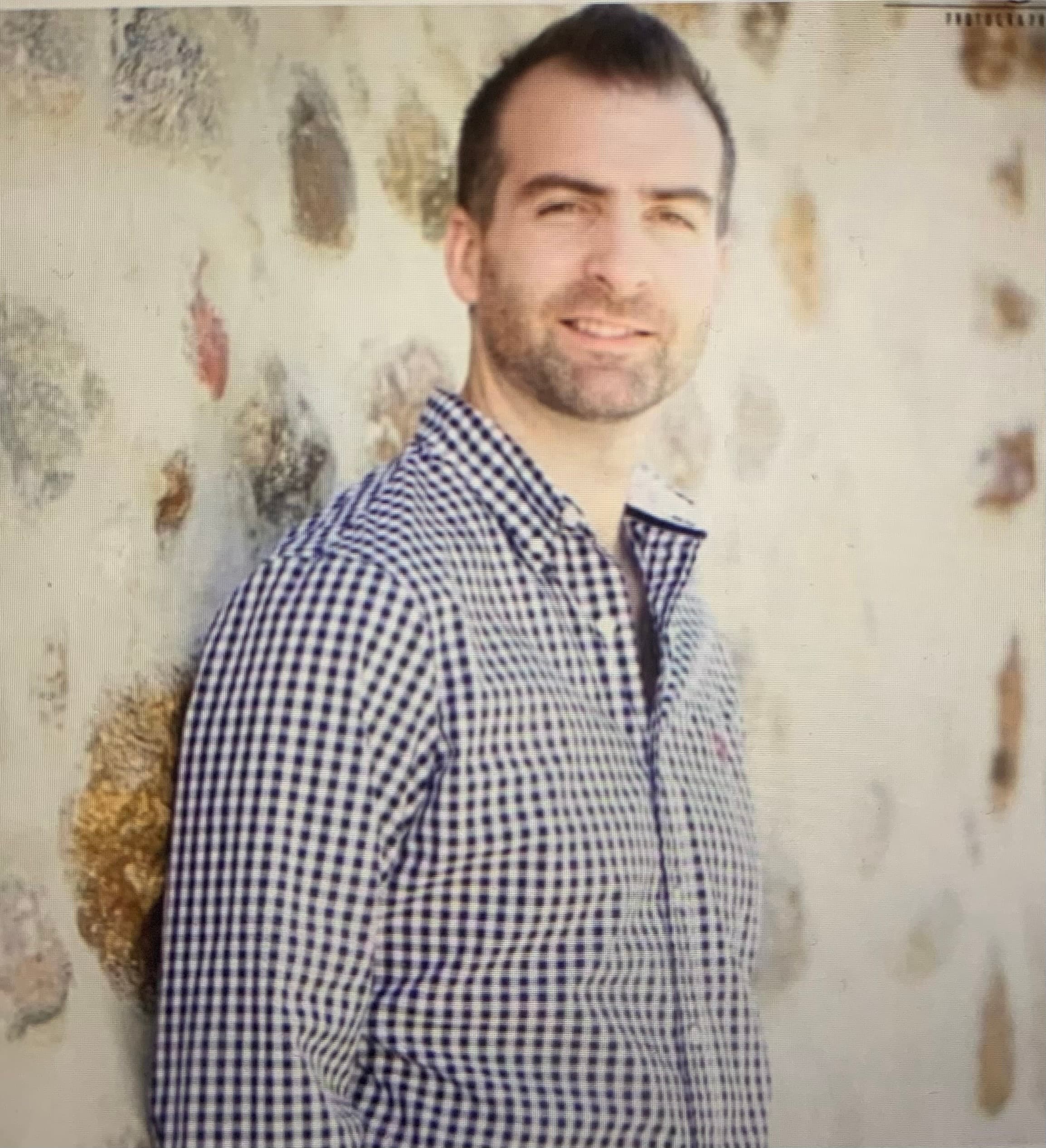 Homme souriant en chemise à carreaux bleue, s'appuyant contre un mur de pierre texturé.
