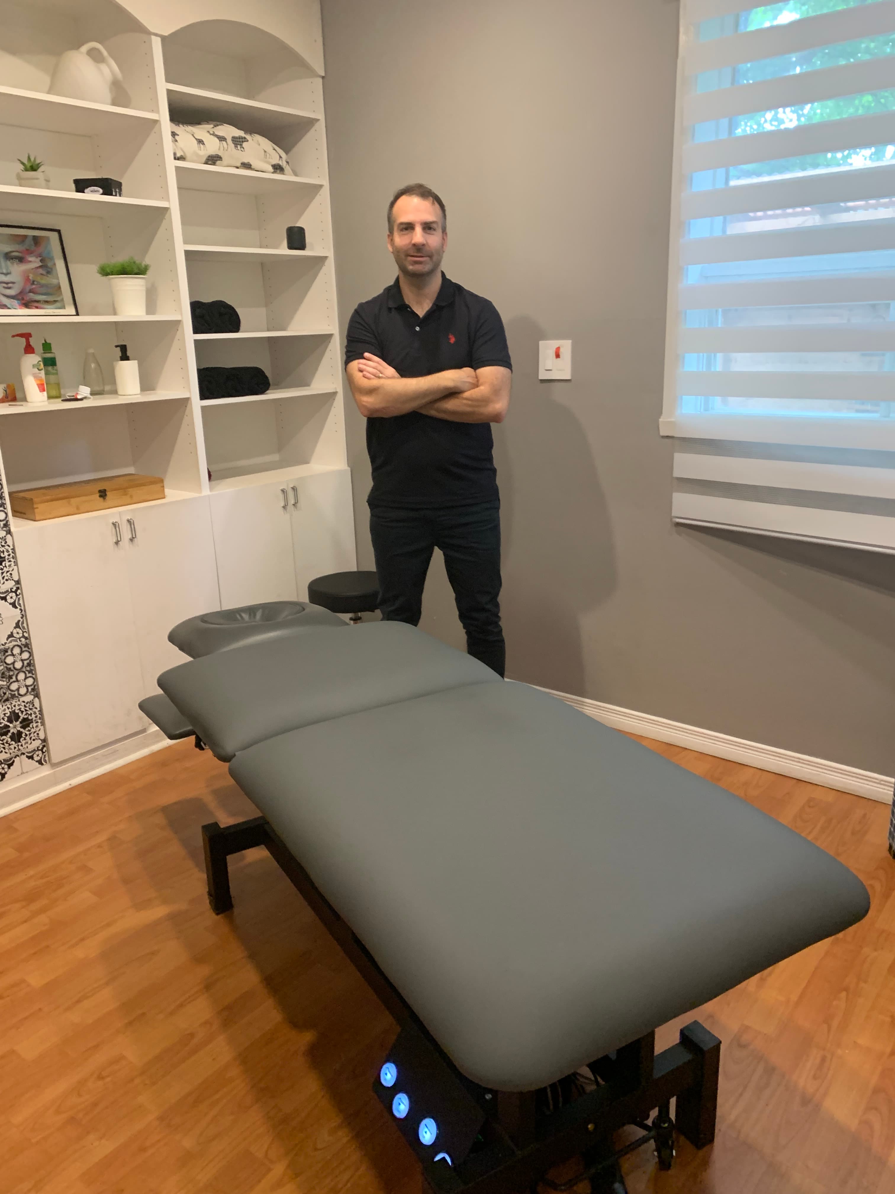 Homme debout les bras croisés derrière une table de massage grise dans son cabinet.
