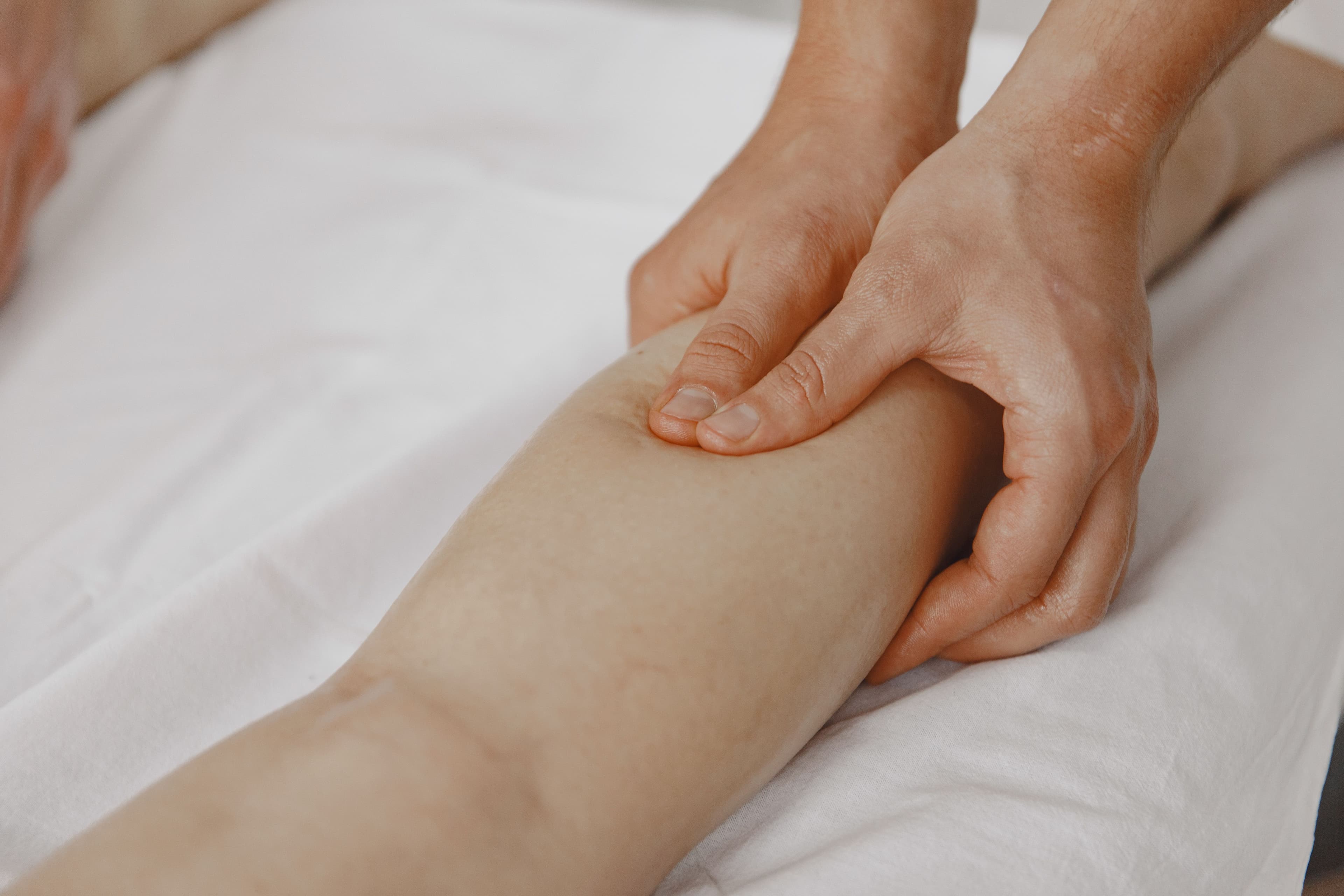 Mains d'un thérapeute massant fermement le mollet d'une personne allongée sur un drap blanc.